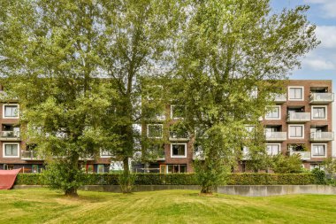 Bereketli yeşil ağaçların çerçevelediği, kentsel yaşam ve doğanın bir karışımını sergileyen modern konut binalarının huzurlu manzarası. Çevreleyen çimenler canlı bir dokunuş sunuyor..