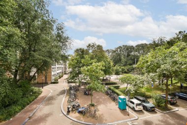 Ağaçlarla kaplı bir park yolu, park edilmiş bisikletler ve arka planda kentsel mimari ile ilgili bir ipucu, rahatlama ve keşifleri davet eden sakin bir sahne..