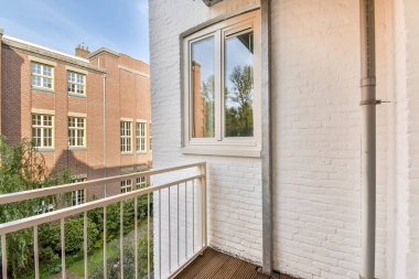 Geniş bir balkon, modern mimari ve doğanın bir karışımını sağlayan tuğla binalar ve yemyeşil alanlarla büyüleyici bir şehir manzarasına bakıyor..