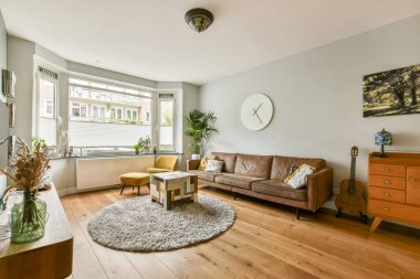 Rahat bir kanepe, dekoratif bitkiler, yuvarlak bir halı ve geniş pencerelerden yayılan doğal ışığın olduğu modern bir oturma odası..