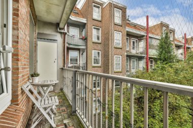 Küçük bir masası ve sandalyeleri olan rahat bir balkon yeşil ve tuğla binalarla çevrili huzurlu bir yerleşim yerine bakıyor..
