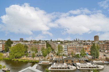 Mavi gökyüzü ve yemyeşil enerjiye zıt, sakin bir kanal boyunca büyüleyici binaların yer aldığı bir şehir manzarası..