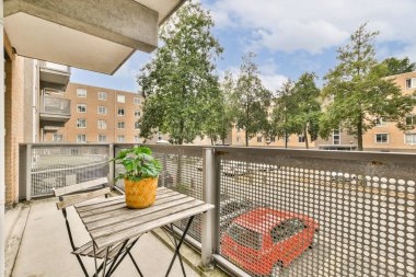 İçinde saksı olan küçük bir masa olan davetkar bir balkon sahnesi, ağaçlarla çevrili kentsel bir çevre ve canlı kırmızı bir araba..