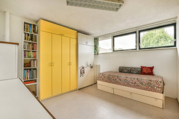 A contemporary bedroom showcasing a cozy bed with a vibrant patterned duvet, bright yellow cabinets, and ample natural light from large windows.
