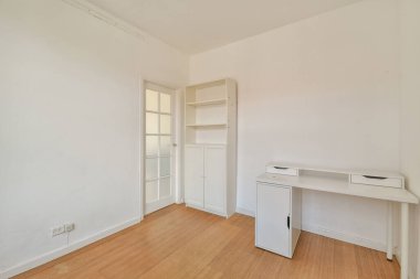 Beyaz bir masa, raf ünitesi ve iş ve rahatlama için sakin bir atmosfer sağlayan davetkar ahşap bir zemin içeren parlak ve basit bir oda..