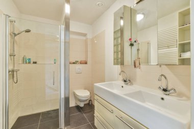 Cam duş muhafazası, minimalist lavabo ve genel estetiği geliştiren nötr renk düzeninde zarif armatürlerin yer aldığı gösterişli modern bir banyo..