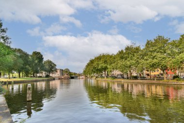 Berrak suları yansıtan sakin bir kanal manzarası. Berrak ağaçlar ve kıyılar boyunca parlak mavi gökyüzünün altında renkli binalar..