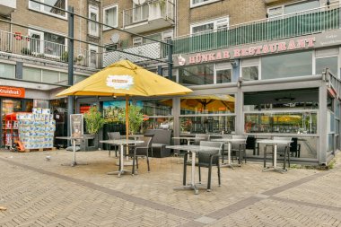 Davetkar bir açık hava restoranında sarı şemsiyelerin altında şehir manzarasıyla çevrili masalar misafirler için sıcak bir yemek atmosferi yaratıyor..