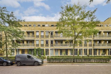 Çerçevesi yemyeşil ağaçlar ve bakımlı sokak boyunca düzgünce biçilmiş çitlerle çevrili, balkonları olan modern bir bina..