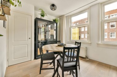 Yuvarlak bir masa, siyah sandalyeler ve geniş pencerelerden içeri ışık sızan parlak, havadar bir atmosfer içeren şık bir yemek salonu..