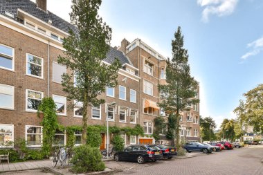 Modern binalar, park edilmiş arabalar ve yeşilliklerle dolu ağaçlarla kaplı bir caddenin canlandırıldığı pitoresk bir şehir manzarası, canlı bir şehir yaşamını yansıtıyor..