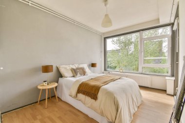 Rahat bir battaniyesi, ahşap sehpaları, doğal ışığa izin veren geniş pencereleri ve sakin bir ortam için minimalist dekorasyonu olan şık bir yatak odası..