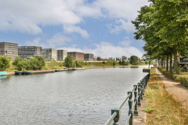 Çağdaş bir şehir manzarasında akan bir nehrin sakin manzarası yeşillik, binalar ve yürüyüş yollarıyla dolu, doğa ve kentsel yaşamın ahenkli bir karışımını gösteriyor..