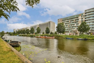 Sudaki çeşitli teknelerin ve nehir boyunca yemyeşil yeşilliklerin yer aldığı, binalar ve ağaçlarla kaplı modern bir sahil manzarası..