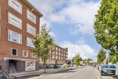 Canlı bir sokak sahnesi ağaçlarla kaplı modern binaları sergiliyor, şehir manzarasını güzelleştiriyor ve yol boyunca bir gezintiye davet ediyor..