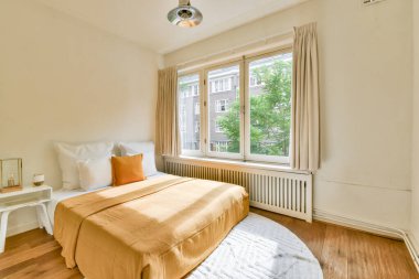 Ilık turuncu bir yatak örtüsü, doğal ışık getiren büyük pencereleri ve rahatlamak için mükemmel sakin bir atmosferi olan huzurlu bir minimalist yatak odası..