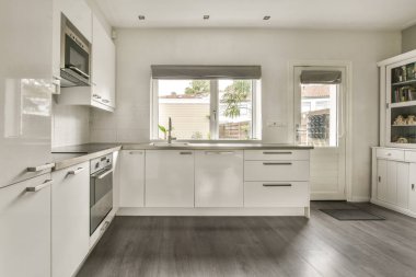 Beyaz dolaplar, paslanmaz çelik aletler ve dışarısı yeşillik manzaralı parlak bir pencere içeren gösterişli ve çağdaş mutfak tasarımı..