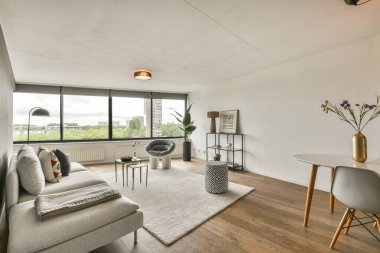 Zarif mobilyaların bulunduğu modern bir oturma odası, geniş pencerelerden geçen doğal ışık, ve rahatlamak veya eğlenmek için mükemmel şık bir atmosfer..