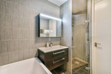 Gösterişli ve çağdaş bir banyo. Minimalist tasarım unsurları sergiliyor. Gri fayanslar, dikdörtgen bir ayna ve depolama depolu şık bir kibir..