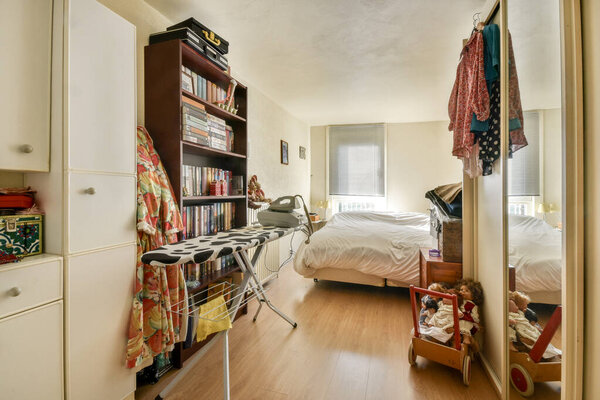 Теплая и уютная спальня с гладильной доской, книжной полкой и уютной кроватью. Декор включает мягкое освещение и разноцветную одежду, драпированную рядом.