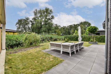 Bereketli yeşilliklerin, manikürlü çimlerin ve parçalı bulutlu gökyüzünün altında modern bahçe mobilyalarının olduğu huzurlu bir arka bahçe sahnesi..