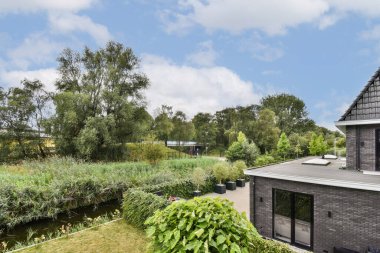 Canlı yeşil bitkiler ve sakin bir su yolu içeren sakin bir manzara, dağınık ağaçlar ve arka planda modern bir bina ile tamamlanmış..