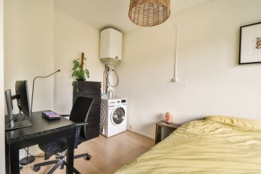 Minimalist dekorlu, sıcak ışıklandırmalı, yatak, masa ve çamaşır makinesinin bulunduğu küçük bir oda rahat bir atmosfer yaratıyor..