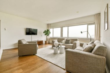 Geniş, modern bir oturma odası, açık renkli duvarlar, rahat koltuklar, bir televizyon ünitesi ve doğal ışığın boşluğu doldurmasına izin veren büyük pencereler..