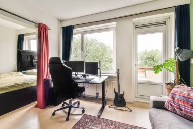 Bilgisayar monitörlü bir masa, rahat bir sandalye, doğal ışık ve gitar ve bitkiler gibi dekorasyon elementlerini rahatlatan modern bir ofis..