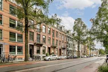 Tarihi binalar, yeşil ağaçlar, park edilmiş bisikletler ve açık gökyüzü altındaki arabaların yer aldığı pitoresk bir şehir sokak sahnesi, şehir hayatı ve doğa karışımı bir şey sergiliyor..