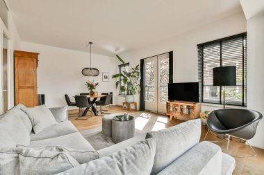 Rahat gri bir kanepe, ahşap mobilyalar ve doğal ışığa izin veren geniş pencereli şık bir oturma odası. Dekoru modern ve davetkar..