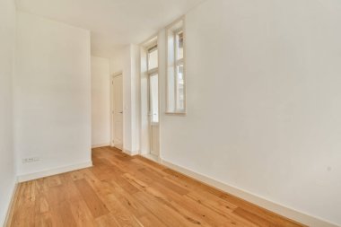 Geniş, minimalist bir iç mekan. Geniş pencerelerden süzülen doğal ışık, ahşap döşemeleri ve temiz beyaz duvarları aydınlatıyor..