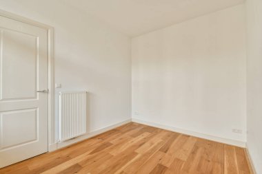 Basit bir kapı ve radyatör ile tamamlanmış, ahşap zemin ve beyaz duvarları olan ferah minimalist bir oda, parlak ve havadar bir atmosfer yaratıyor..