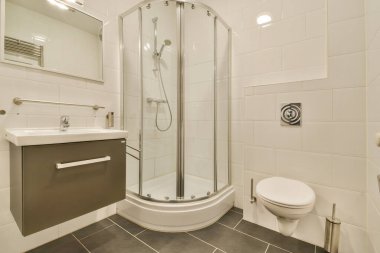 Parlak ve çağdaş bir banyo. İçinde cam duş muhafazası, modern eşyalar ve temiz, beyaz fayanslar var. Alan işlevsellik ve tarz için tasarlanmıştır..
