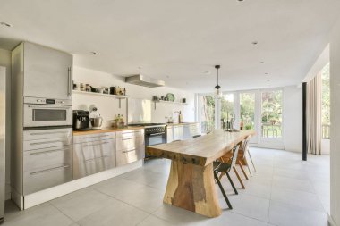 Zarif paslanmaz çelik aletler, ahşap bir yemek masası ve doğal ışık getiren büyük pencereleri olan güzel tasarlanmış modern bir mutfak..