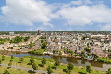 Evleri, yeşillikleri ve kıvrımlı bir su yolu olan canlı bir şehir alanını gösteren geniş bir hava görüntüsü. Sahne, doğa ve şehir hayatının karışımını vurguluyor..