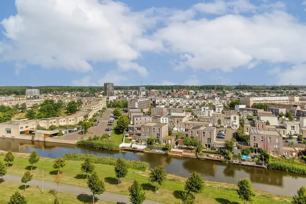 Evleri, yeşillikleri ve kıvrımlı bir su yolu olan canlı bir şehir alanını gösteren geniş bir hava görüntüsü. Sahne, doğa ve şehir hayatının karışımını vurguluyor..