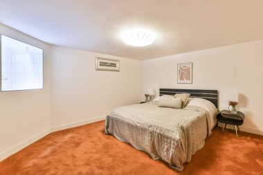 Geniş ve rahat bir yatak odası, gri çarşaflarla süslenmiş rahat bir yatak odası, şık bir aydınlatma armatürü ve yumuşak turuncu halı. Doğal ışık pencereden içeri sızar..