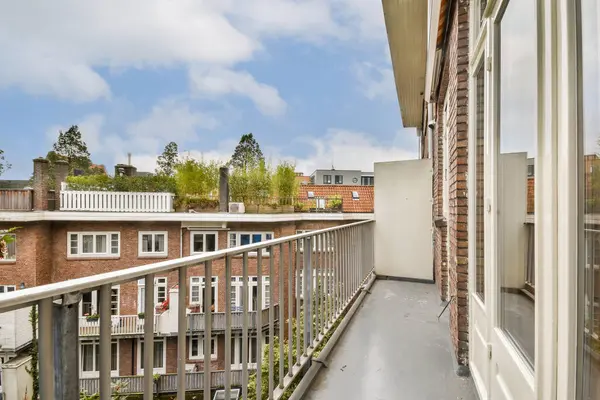 Sakin bir balkon manzarası yeşillik ve şehir mimarisinin bir karışımını sergiliyor. Sahne, açık bir gökyüzünün arka planına karşı yerleşim binalarını ön plana çıkarıyor.