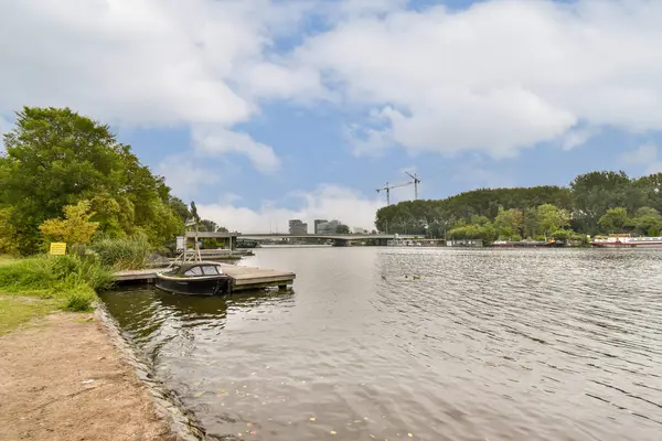Sakin bir su yolu, küçük bir tekne ve parçalı bulutlu bir gökyüzü altında çevreleyen yeşillik içeren pitoresk bir nehir sahnesi.