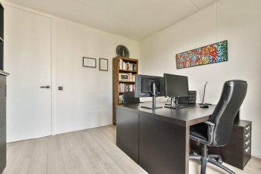 Ahşap bir masa, bilgisayar kurulumu, kitap rafları ve sanatsal duvar dekorunun yer aldığı modern bir ofis üretken bir çalışma alanı ortamı yaratıyor..