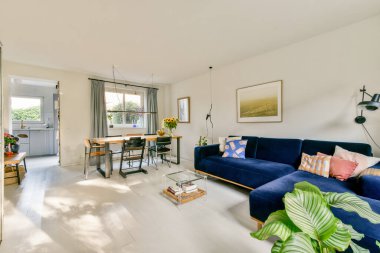 Mavi bir kanepe, ahşap bir yemek masası ve pencerelerden süzülen parlak güneş ışığı olan şık bir oturma odası sıcak, davetkar bir atmosfer yaratıyor..