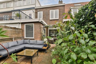 Güzel bahçedeki yemyeşil bitkilerle çevrili şık gri bir kanepe ve ahşap bir sehpa içeren rahat bir açık hava..