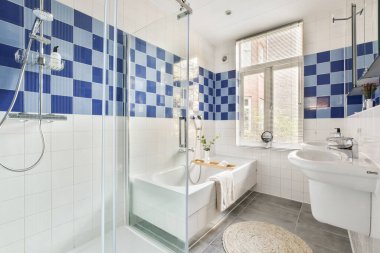 İyi tasarlanmış, mavi ve beyaz fayanslı duvarları olan modern bir banyo, cam bir duş ve camdan gelen doğal ışıkla tamamlanmış bir küvet..