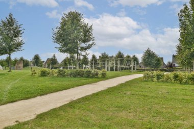 Dolambaçlı bir patika, canlı bir yeşillik ve bulutlu gökyüzünün altındaki ağaçların arasında oyun ekipmanlarıyla dolu huzurlu bir park ziyaretçileri rahatlamaya ve keyif almaya davet ediyor..