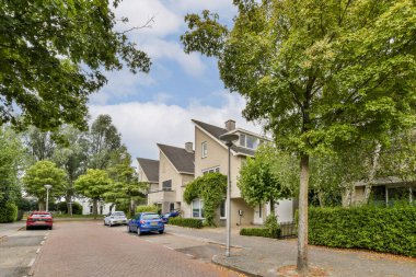 Modern evler, yemyeşil ağaçlar ve bulutlu bir gökyüzünün altında park edilmiş arabalarla dolu sakin bir banliyö caddesi huzurlu bir mahalle atmosferi yaratıyor..