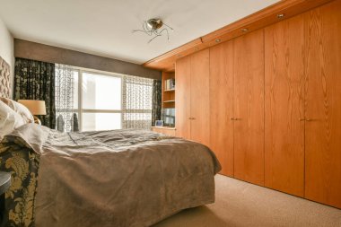 Modern bir yatak odası, ahşap dolaplar, rahat bir yatak, ve zarif perdeler, dinlenmek için sıcak ve davetkar bir atmosfer yaratıyor..