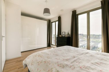 Rahat, modern bir yatak odası çiçek desenli bir yorgan, geniş pencereler, doğal ışık ve rahatlatıcı bir atmosfer için zarif perdeler..