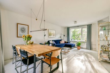 Ahşap yemek masası, mavi kanepe ve doğal ışık ve yeşillik getiren büyük pencereli şık bir yaşam alanı..