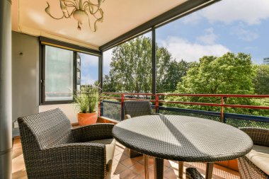Rahat dokuma mobilyaların olduğu şık bir balkon, ağaçların manzarası, ve rahatlama ve toplanma için mükemmel mavi bir gökyüzü..